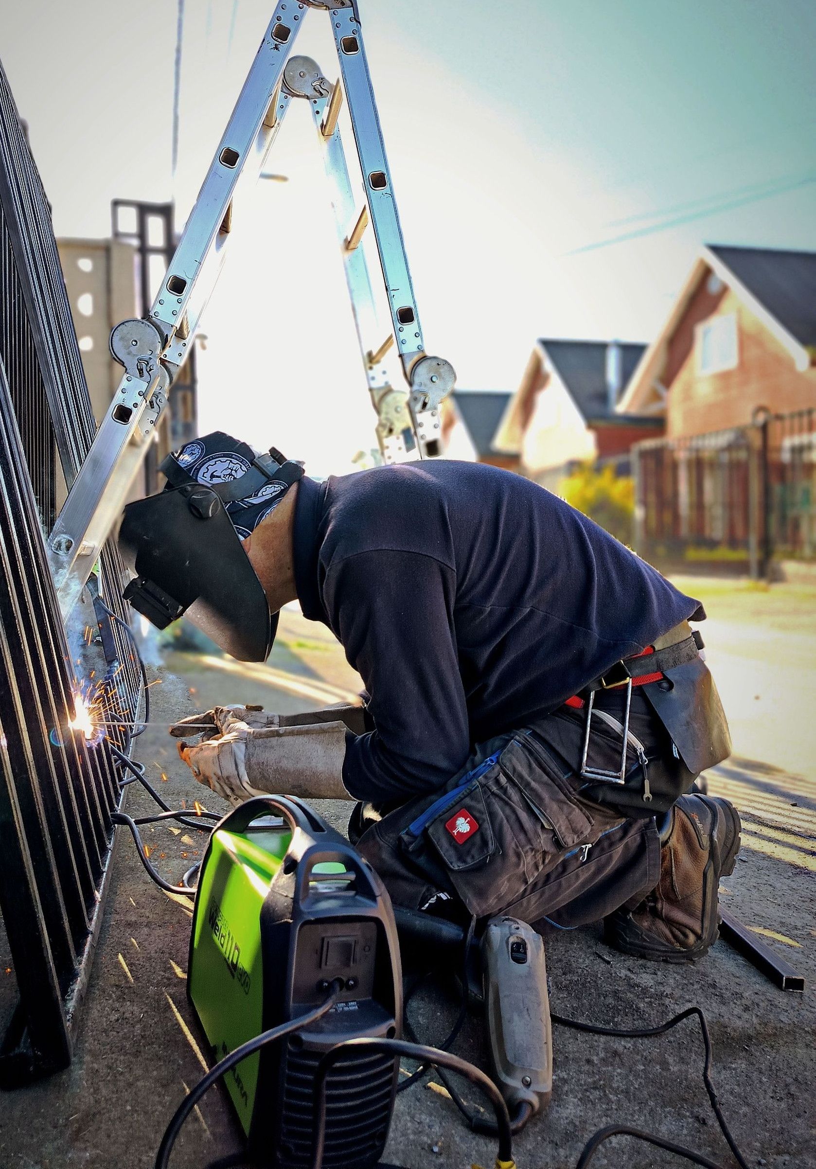 Welder wearing protective gear working on a metal fence during the day with welding equipment.